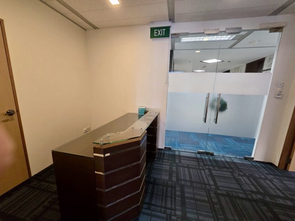 A reception desk is positioned near a glass door with frosted panels, leading to a corridor with blue patterned carpet and an illuminated exit sign above the door.
