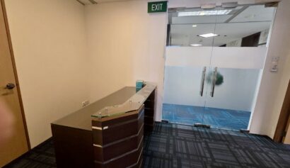 A reception desk is positioned near a glass door with frosted panels, leading to a corridor with blue patterned carpet and an illuminated exit sign above the door.