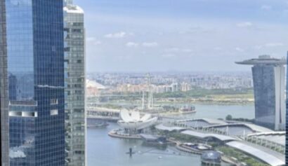 Aerial view of tall glass skyscrapers, waterfront, and modern city architecture with blue sky and scattered clouds.
