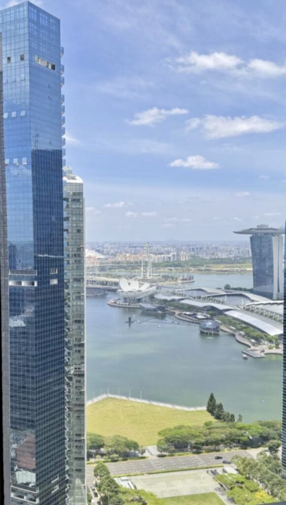 Aerial view of tall glass skyscrapers, waterfront, and modern city architecture with blue sky and scattered clouds.