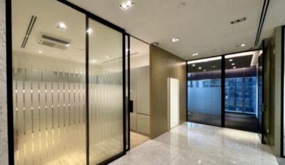 Modern office interior with glass walls, frosted glass partitions, tiled floors, and ceiling lights, featuring a corridor leading to a separate office space.