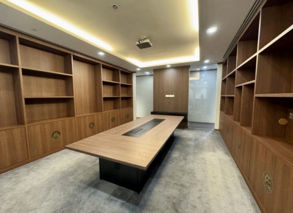 Modern conference room with built-in wooden shelves, a long table at the centre, recessed lighting, and carpeted floor.