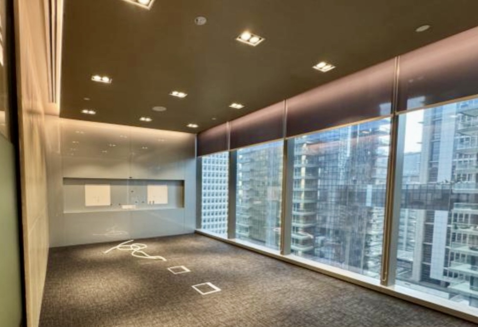 Modern empty office space with carpeted floor, ceiling lights, built-in wall cabinetry, and large floor-to-ceiling windows overlooking a cityscape of tall buildings.
