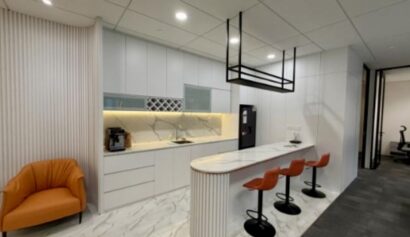 Modern office kitchenette with white cupboards, marble worktop, three orange barstools, an orange armchair, and a coffee machine on a marble floor.