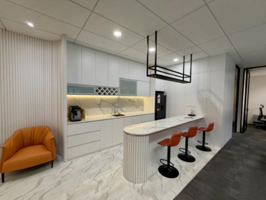 Modern office kitchenette with white cupboards, marble worktop, three orange barstools, an orange armchair, and a coffee machine on a marble floor.