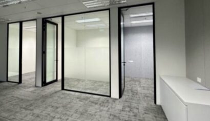 Empty modern office space with glass-walled rooms, grey carpet, white walls, and a white storage cupboard against the right wall.