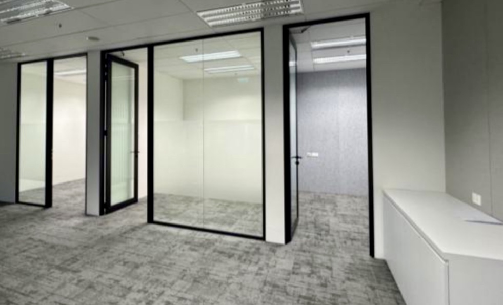 Empty modern office space with glass-walled rooms, grey carpet, white walls, and a white storage cupboard against the right wall.