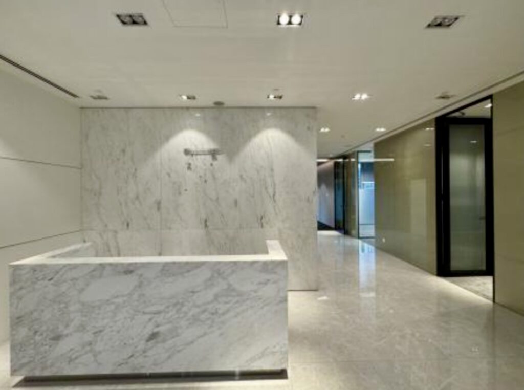 A modern office lobby with marble floors, a marble reception desk, and glass-walled offices along the corridor.