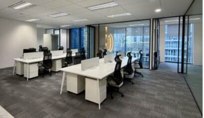 Modern office space with several white desks and black chairs arranged in rows, glass walls, and large windows revealing city buildings outside.