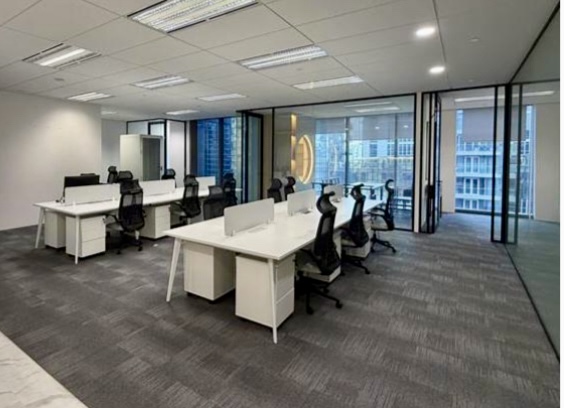 Modern office space with several white desks and black chairs arranged in rows, glass walls, and large windows revealing city buildings outside.