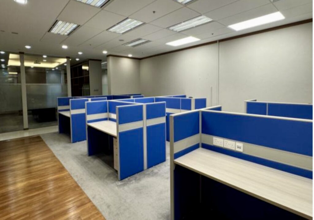 An empty office space with several blue cubicles, overhead fluorescent lighting, and a combination of carpeted and wooden flooring.