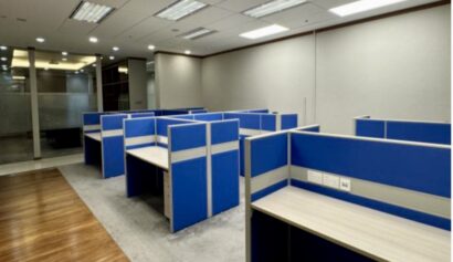 An empty office space with several blue cubicles, overhead fluorescent lighting, and a combination of carpeted and wooden flooring.