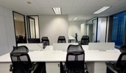 Modern office space with several empty desks, black mesh chairs, and glass-walled meeting rooms in the background.