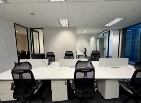 Modern office space with several empty desks, black mesh chairs, and glass-walled meeting rooms in the background.
