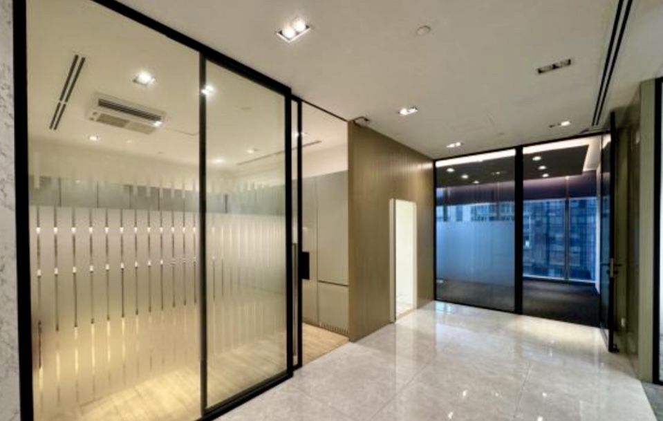 Modern office interior with glass walls, frosted glass partitions, tiled floors, and ceiling lights, featuring a corridor leading to a separate office space.