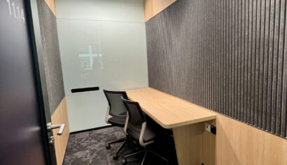 Small office room with two black chairs, a wooden desk, grey patterned carpet, and walls featuring vertical black panels and light wood accents.