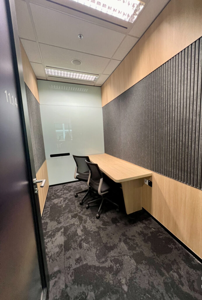 Small office room with two black chairs, a wooden desk, grey patterned carpet, and walls featuring vertical black panels and light wood accents.