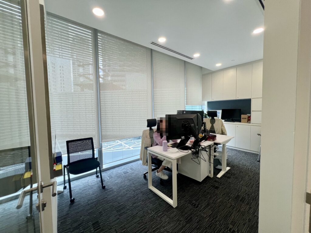 Modern office with two people working at desks with computers, large windows with blinds, white cupboards, and a black chair near the door.