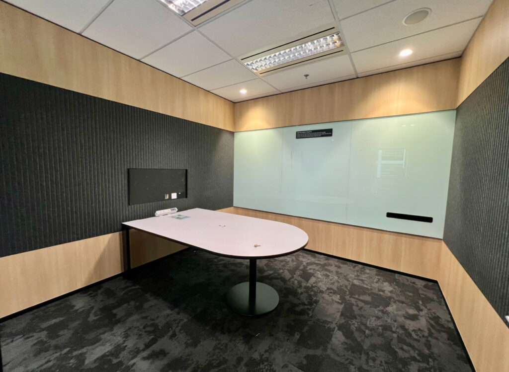 Small modern meeting room with a white table, black feature wall, wood panelling, and a large whiteboard on one wall. Ceiling lights are on, and there are no chairs present.
