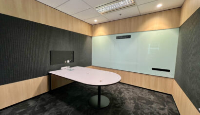 Small modern meeting room with a white table, black feature wall, wood panelling, and a large whiteboard on one wall. Ceiling lights are on, and there are no chairs present.