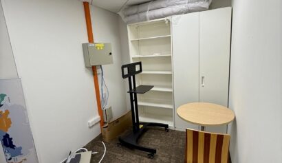 Small office room with a brown carpet, a round table and striped chair, a white shelving unit, a cupboard, a wall-mounted control panel, and some loose cables and equipment on the floor.