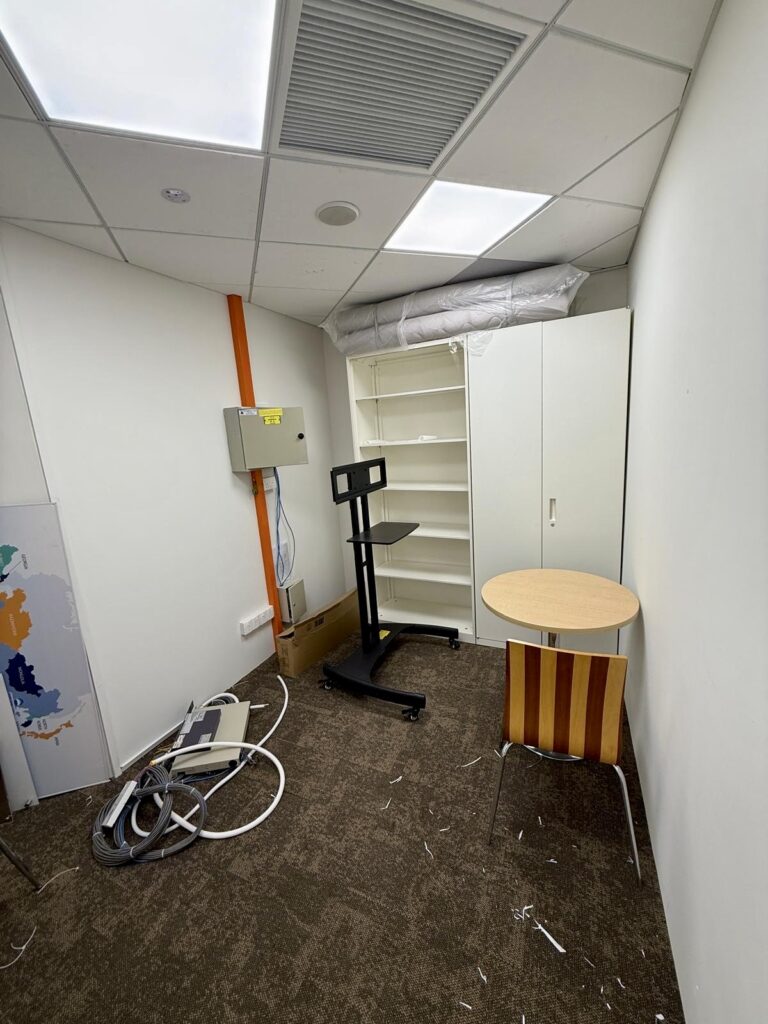 Small office room with a brown carpet, a round table and striped chair, a white shelving unit, a cupboard, a wall-mounted control panel, and some loose cables and equipment on the floor.