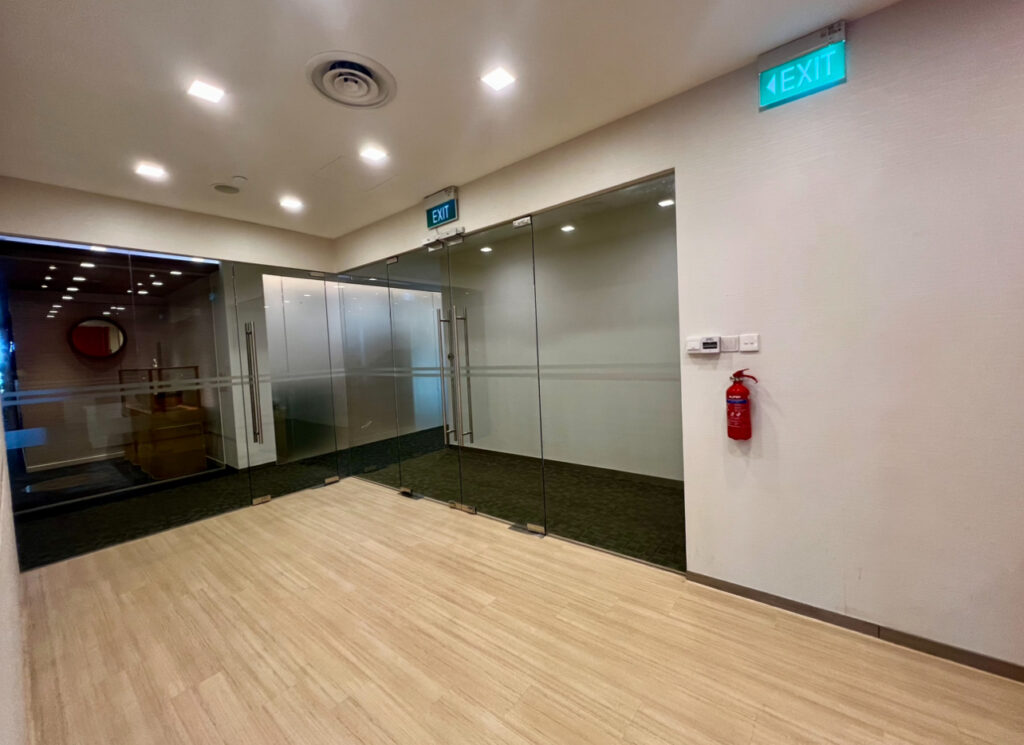 Empty corridor with glass walls, wooden flooring, a visible fire extinguisher on the right wall, and illuminated exit signs above glass doors.