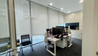 Modern office with two people working at desks with computers, large windows with blinds, white cupboards, and a black chair near the door.