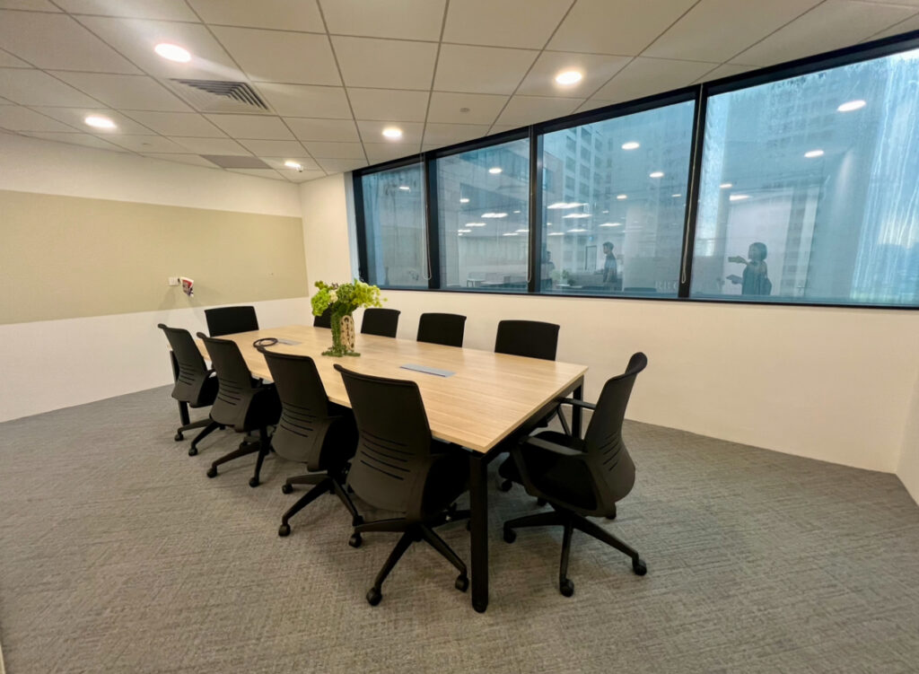 A modern meeting room with a rectangular table, eight black chairs, a vase of flowers, and large windows overlooking office buildings.