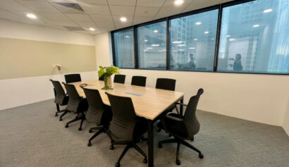 A modern meeting room with a rectangular table, eight black chairs, a vase of flowers, and large windows overlooking office buildings.