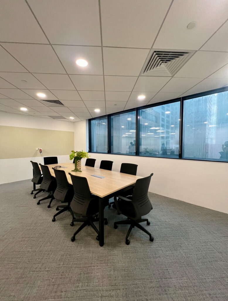 A modern conference room with a long table, black chairs, a plant centrepiece, and large windows showing an exterior view.