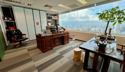 Spacious corner office with wooden desk, bookshelves, a large window overlooking a cityscape, and a potted plant near a meeting table.