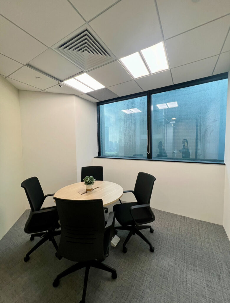 A small office room with a round table, four black chairs, a potted plant, and large windows showing reflections of people.