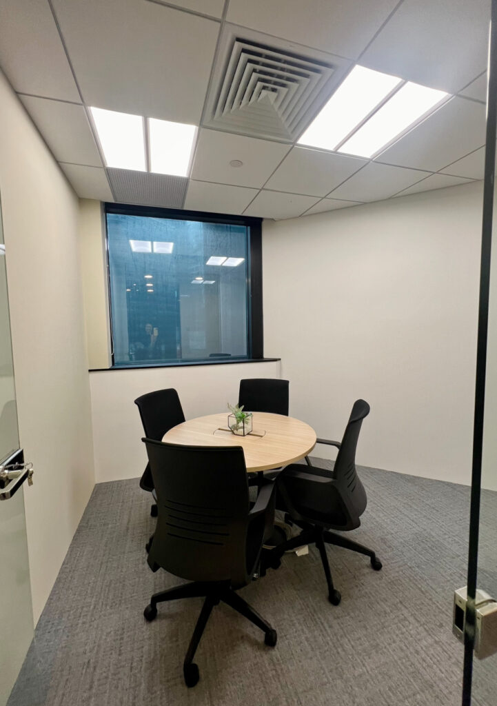 Small meeting room with four black chairs around a round wooden table, carpeted floor, large window, and neutral walls.