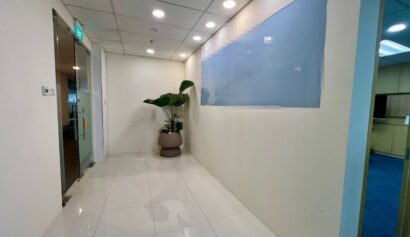 A clean, well-lit office corridor with white walls, tiled floor, a glass door on the left, a potted plant, and a large frosted glass panel on the right wall.
