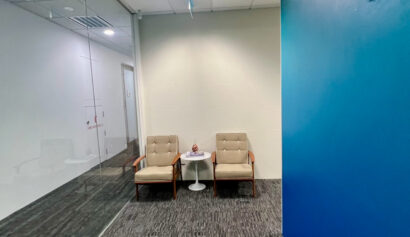 Two beige armchairs and a small white round table with a decorative item sit against a white wall in a modern office corridor with blue accents and glass partitions.