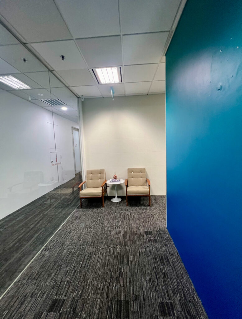 Two beige armchairs and a small white round table with a decorative item sit against a white wall in a modern office corridor with blue accents and glass partitions.