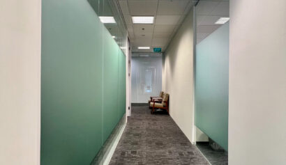 A modern office corridor with glass walls, grey carpet, and a brown armchair at the end near a door with a green exit sign above it.