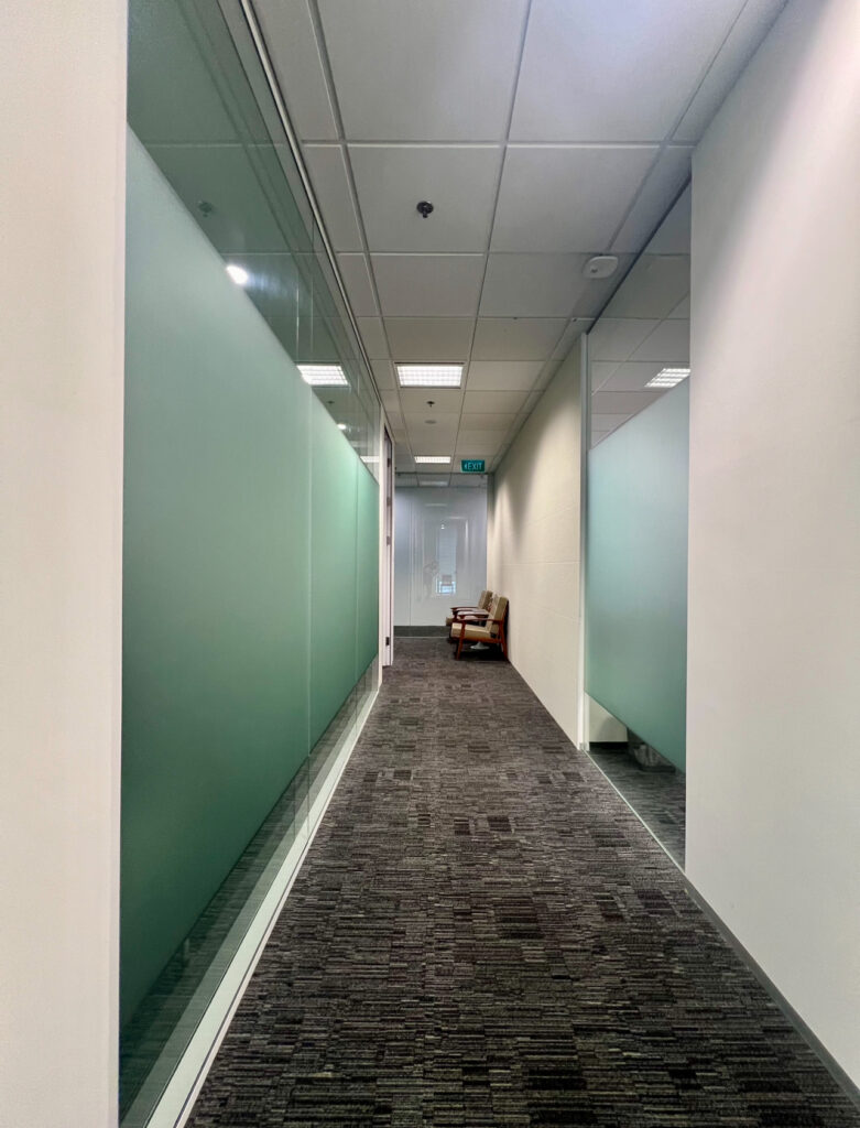A modern office corridor with glass walls, grey carpet, and a brown armchair at the end near a door with a green exit sign above it.