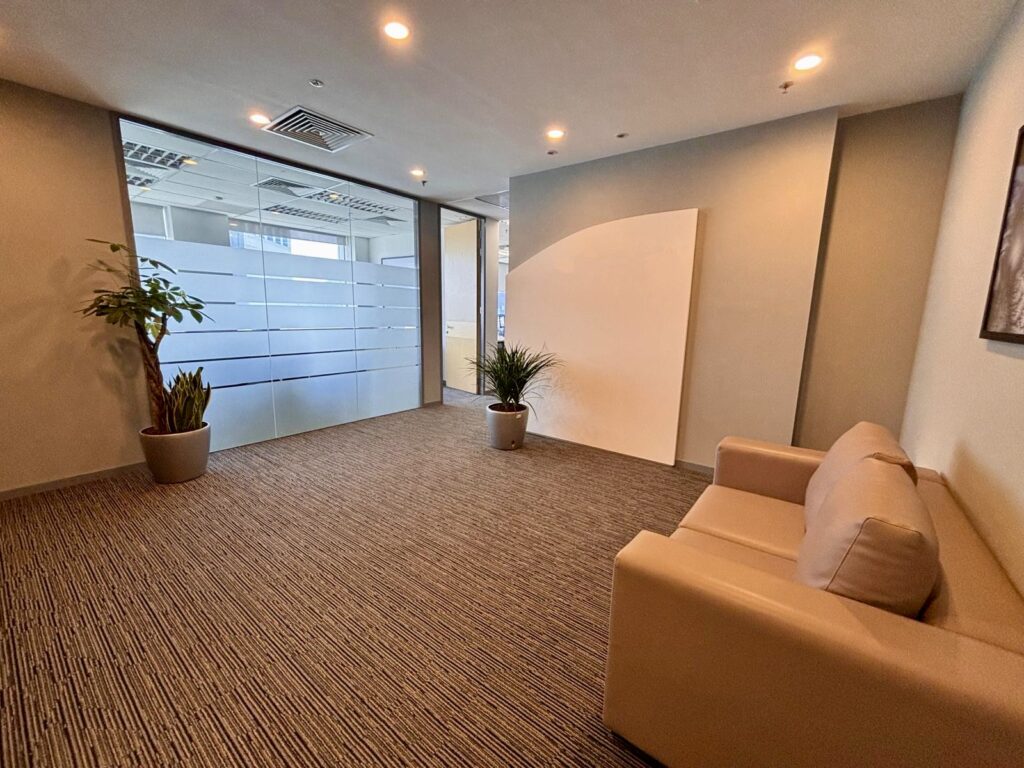 A modern office waiting area with a beige sofa, two potted plants, carpeted floor, and frosted glass partition walls.