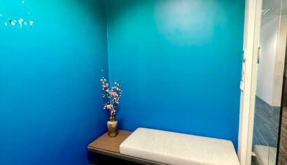 A small room with teal walls, a grey cushioned bench, and a vase with decorative branches on a wooden surface.