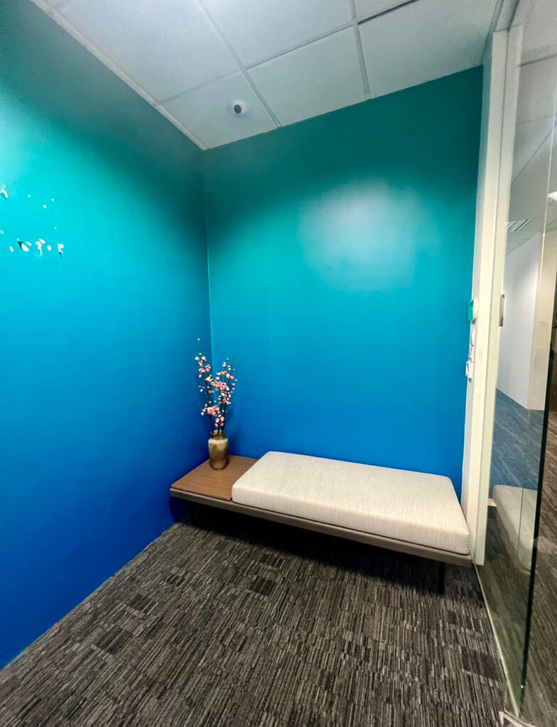 A small room with teal walls, a grey cushioned bench, and a vase with decorative branches on a wooden surface.