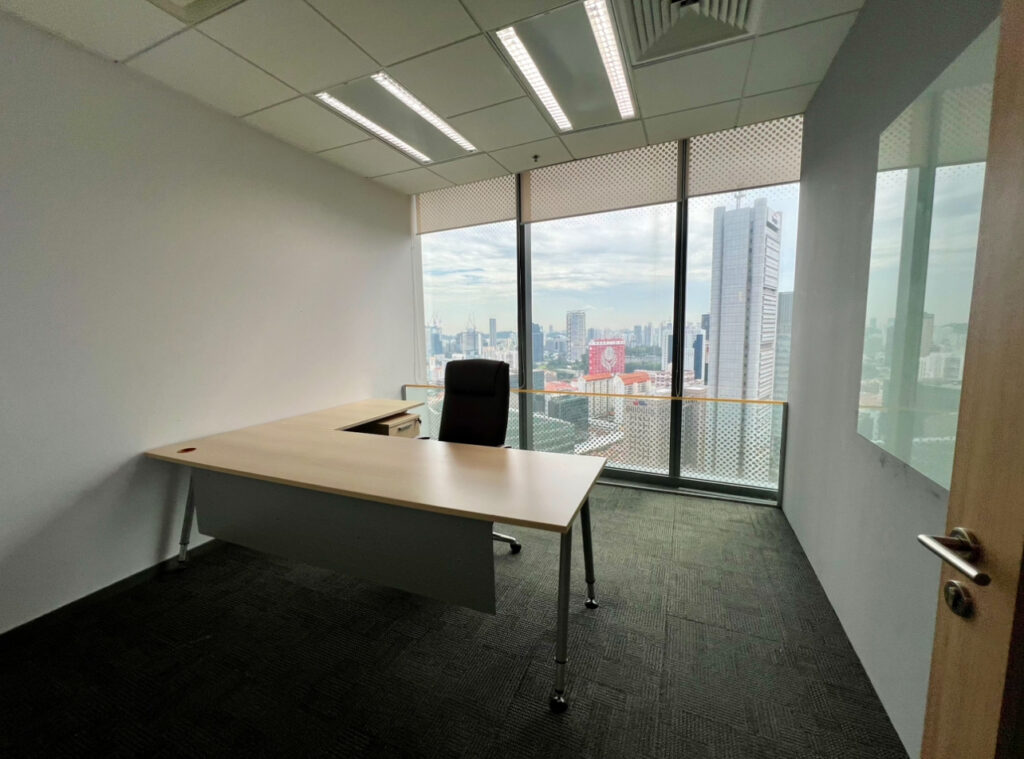 A modern office with a desk, chair, and large windows overlooking a cityscape on a cloudy day. The room has white walls and dark carpet.