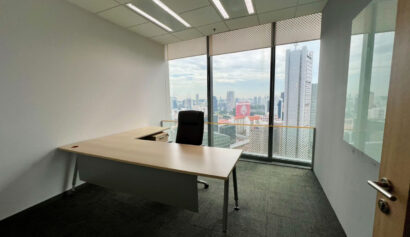 A modern office with a desk, chair, and large windows overlooking a cityscape on a cloudy day. The room has white walls and dark carpet.