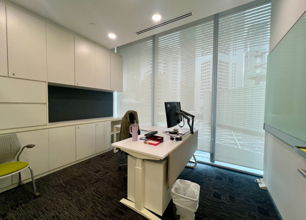 Modern office with white cupboards, a desk with computer monitor, chair, water bottle, and snacks. Large windows with blinds show city buildings outside.