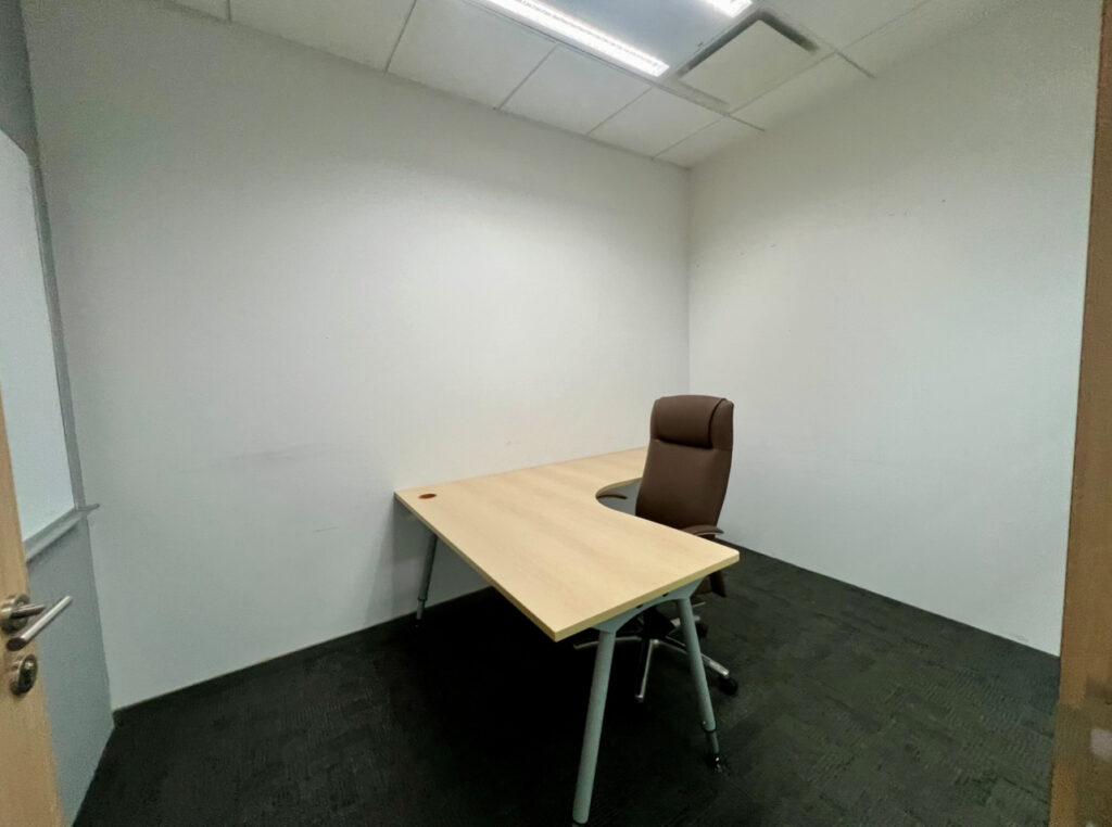 Small office room with a single empty wooden desk and a brown office chair, white walls, and a closed door partially visible.