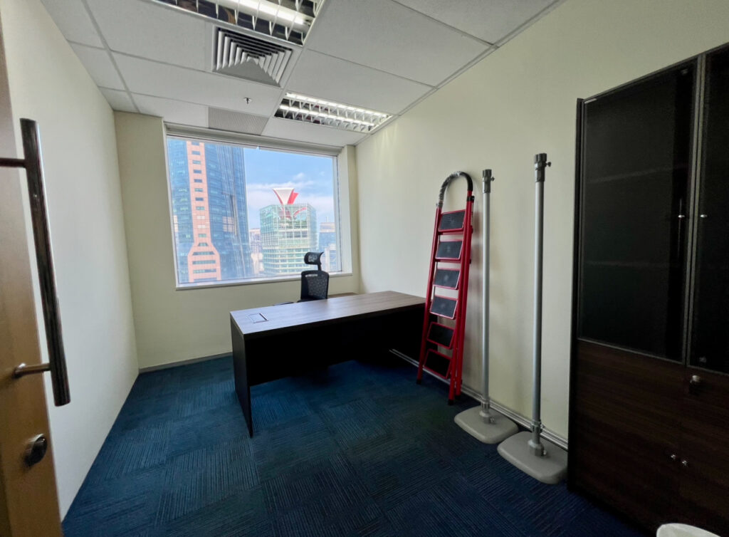 Small office with a desk, chair, red ladder, coat racks, and cupboard. Large window shows city buildings outside. Blue carpet and white walls.