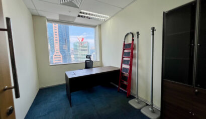 Small office with a desk, chair, red ladder, coat racks, and cupboard. Large window shows city buildings outside. Blue carpet and white walls.