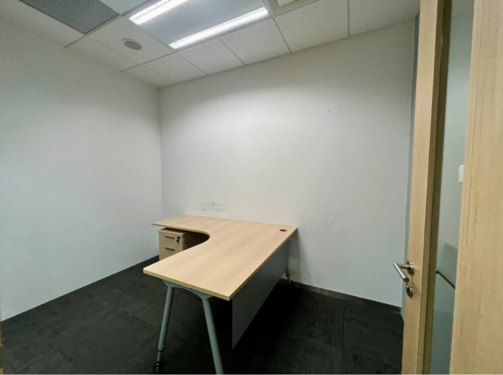 Small, empty office room with a light-coloured L-shaped desk, single drawer unit, plain white walls, and black carpet flooring. Overhead fluorescent lights are switched on.