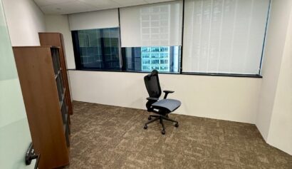 Empty office room with brown carpet, a black office chair, a wooden bookcase, large windows with blinds, and a power strip on the floor.